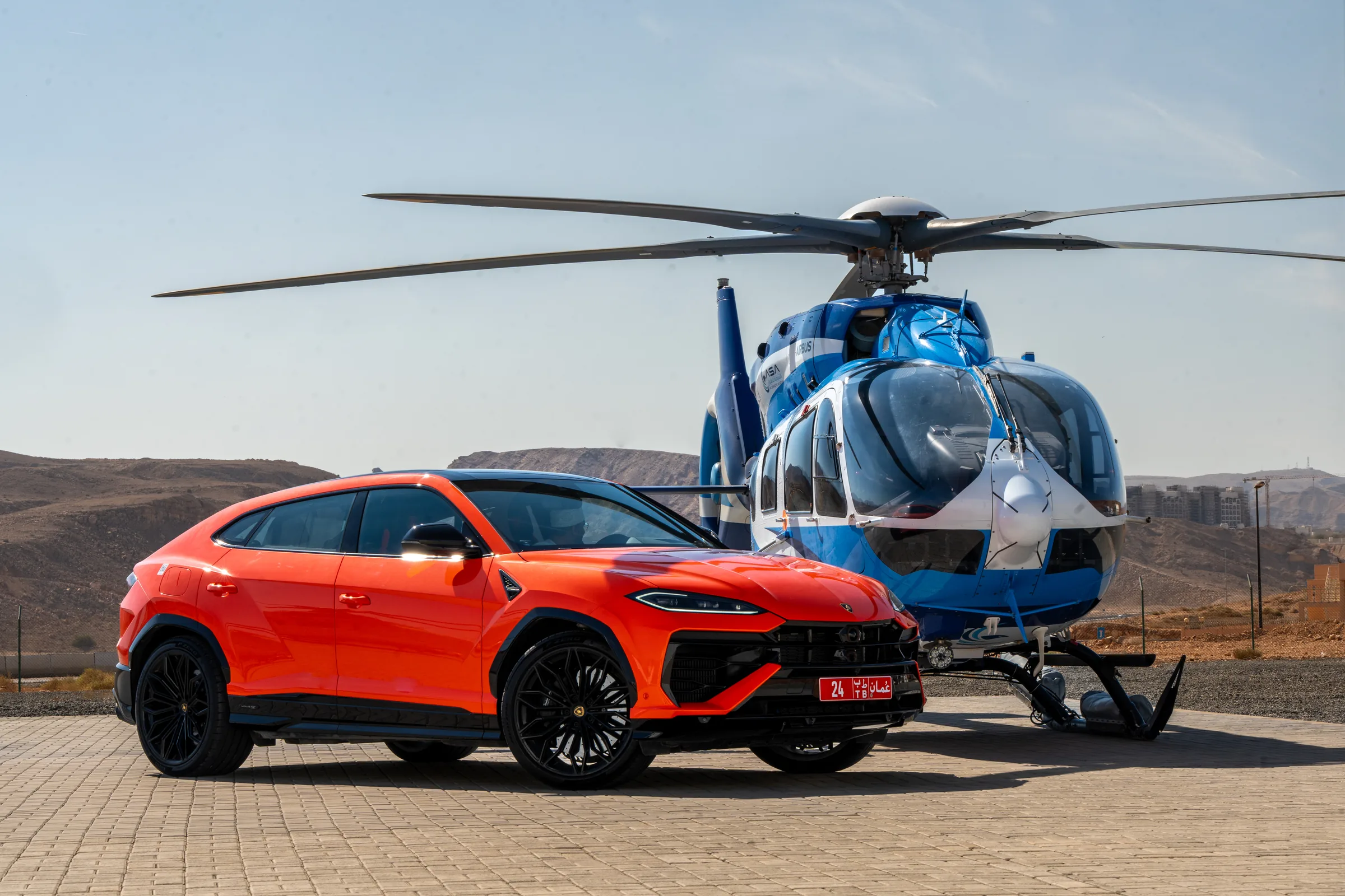 Orange Lamborghini Urus parked beside a helicopter with Oman mountains backdrop